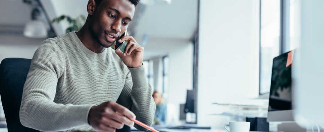 Man sitting at desk talking on phone and looking at desk pointing with a pencil